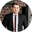 Man in a dark suit and red tie smiling in front of a blurred auditorium background.