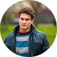 Young man with brown hair wearing a black leather jacket and striped sweater outdoors.